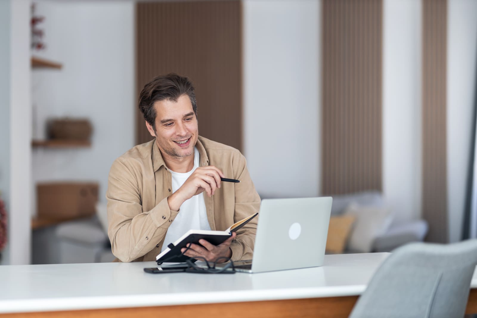 Hombre joven sonriendo mientras toma notas frente a su laptop en casa durante una asesoría en línea