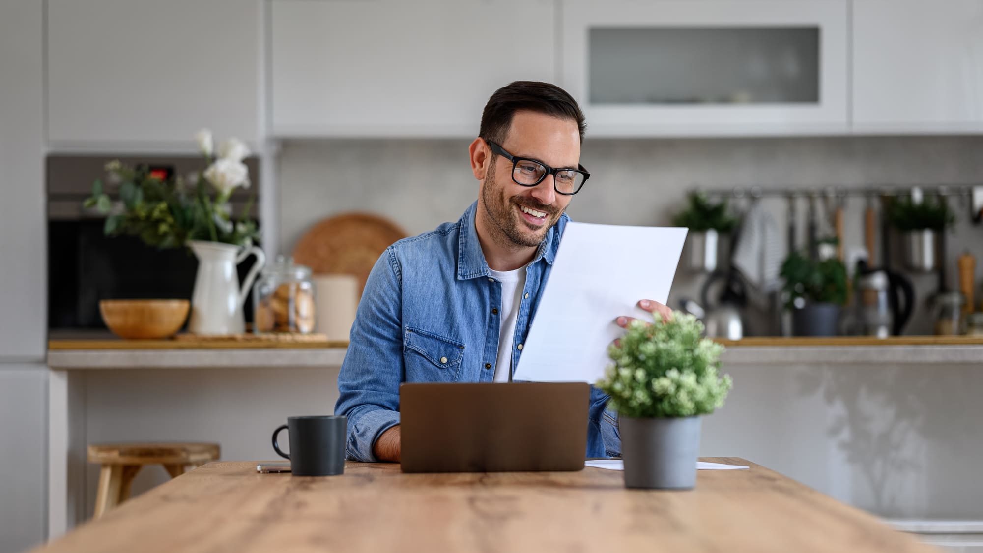 Hombre sonriendo mientras revisa un documento fiscal junto a su laptop en la cocina de su casa